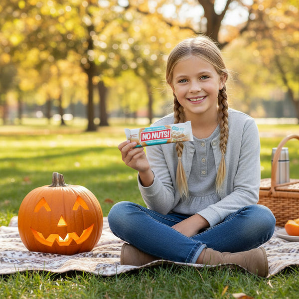 Smiling girl at picnic holding No Nuts nut-free snack bar beside a carved pumpkin outdoors