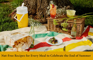 Nut-free picnic with snacks, drinks, and a wicker basket on a colorful blanket outdoors.