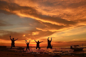 Four people jumping on the beach at sunset with dramatic orange sky and boats in the background.