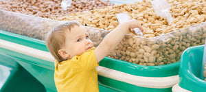 Toddler in yellow shirt reaching for nuts in bulk bin, highlighting nut allergy risk for babies.
