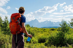 Hiker with orange backpack and water bottle in a green field facing mountains, outdoors adventure