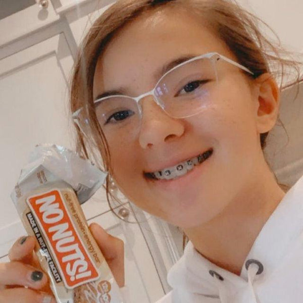Smiling girl with braces holds a No Nuts nut-free snack bar in a bright kitchen.