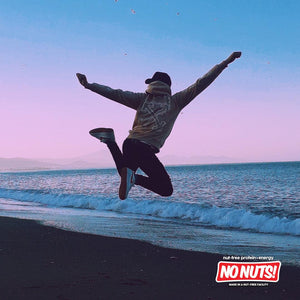 Person joyfully jumping on a beach at sunrise, No Nuts nut-free snack bars logo in corner.