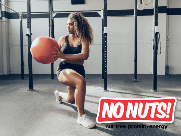 Woman working out with medicine ball in gym, No Nuts nut-free protein energy branding displayed.