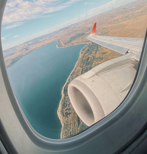 View from airplane window showing wing, engine, lake, and coastline under blue sky.
