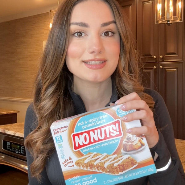 Woman in kitchen holding No Nuts cinnamon roll nut-free protein bars box